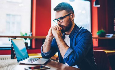 Düşünceli erkek programcı, günlük kıyafetlerini giyer ve şık bir kafede dizüstü bilgisayar ve akıllı telefonla çalışırken işi düşünür.