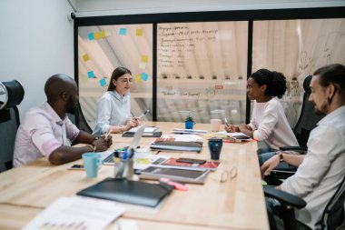 İyimser, gözlüklü, kalemle gülümseyen genç bir kadın iş arkadaşlarıyla birlikte modern ofis odasındaki masada oturan stratejiyi açıklayan bir proje yaratıyor. 