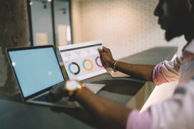 Ofis masasında otururken, diyagramdan gelen verileri analiz eden ve bilgiyi bilgisayara kopyalayan konsantre Afro-Amerikan işçilerin ekin kenar görüntüsü