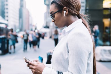 Çağdaş şehrin bulanık arka planında yürürken şık kıyafetli ve gözlüklü siyahi kadınların akıllı telefon kullanışının yan görüntüsü