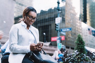 Sosyal medyada gülümseyen ve mesajlaşan gözlüklü mutlu siyahi kadın şehrin bulanık arka planında oturup müzik dinliyor.