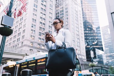 Düşük açılı başarılı Afro-Amerikan kadın profesyonel kulaklık takıyor kaldırımda duruyor ve arka plandaki akıllı telefondan mesajlaşıyor gökdelenler ve ABD bayrakları ile