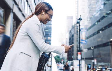 Gözlüklü, kablosuz kulaklıklı, ekranda odaklanan ve caddede bulanık şehir manzarasına karşı durarak akıllı telefonla etkileşim kuran düşünceli Afrikalı Amerikalı kadın.