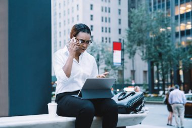 Sinirli Afro-Amerikan bayan çalışanın şehir merkezindeki bankta tabletle otururken cep telefonuyla gergin bir şekilde konuşması.