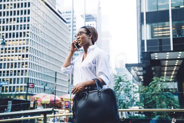 From below of African American woman in formal clothes and eyeglasses with big black handbag talking on smartphone surprised with good news while standing at downtown area with modern high buildings
