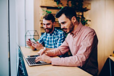 Resmi gömlekli, sakallı, siyah erkek girişimcinin şık bir kafede vakit geçirirken dizüstü bilgisayarla çalışmasının yan görüntüsü. 