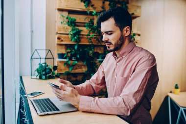 Tarz sahibi kafede dizüstü bilgisayarla çalışırken resmi tişörtle akıllı telefona göz atan meşgul erkek serbest çalışanın yan görüntüsü 