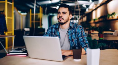 Düşünceli bir erkek, gündelik giysiler içinde bir fincan kahve içiyor. Kafede dizüstü bilgisayar kullanıyor ve gözlerini başka yöne çeviriyor.