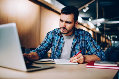 Gündelik kıyafet giymiş yakışıklı bir erkek, kafedeki masada dizüstü bilgisayarla proje üzerinde çalışıyor.