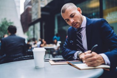 Kibar klas iş adamı, sokak kafesinde masada oturup defter ve tabletle çalışarak tek kullanımlık bardakta içki içiyor.
