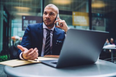 Akıllı takım elbiseli zeki bir işadamı iş merkezindeki yuvarlak masada dizüstü bilgisayarla konuşurken akıllı telefon ve el kol hareketi yapıyor.