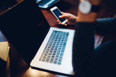 Koyu kütüphanedeki lambanın ışığında açık dizüstü bilgisayarda oturan akıllı telefonlara göz atan kırpıcı.