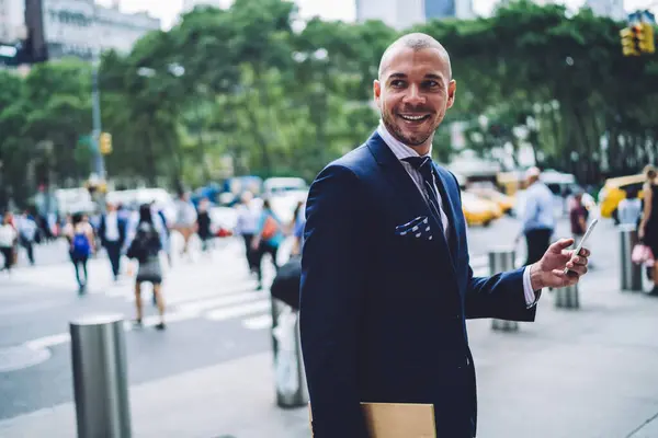 Happy businessman in navy blue suit standing with folder and messaging on smartphone while looking away on blurred background with people walking on crosswalk