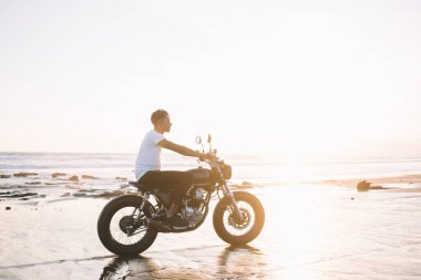 Gün doğumunda boş deniz kıyısında motosiklet süren günlük beyaz tişörtlü ve kot pantolonlu etnik erkek turistin yan görüntüsü