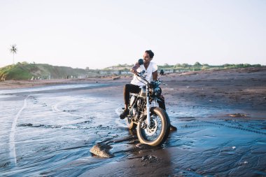 Normal kıyafetlerle gülümseyen yetişkin bir erkek, gidonu tutuyor ve motosikletin üzerinde otururken dizine yaslanıyor ve kırsalın bulanık arka planında ıslak kumsala bakıyor.