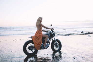 Havalı, sarı saçlı, uzun turuncu elbiseli bir kadının ıslak kumlu sahilde motosiklete binip kameraya bakışının yan görüntüsü. 