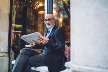 Sakallı, kahve fincanı bardaklı ve takım elbiseli yakışıklı adam beyaz duvarın yanında oturmuş gazete okuyor.