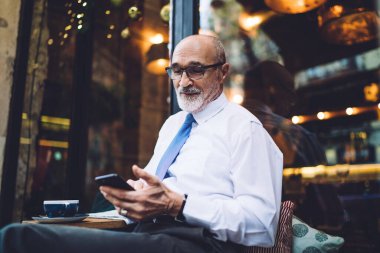 Gözlüklü, beyaz gömlekli, gülümseyen ve cadde kafesinin penceresinde oturan ve akıllı telefondan mesaj atan kıdemli yakışıklı adam.