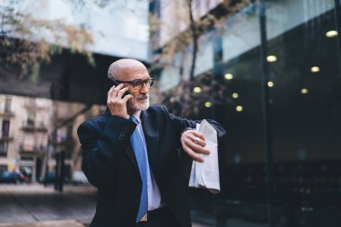 Kendine güvenen, zarif giyinmiş, gözlüklü, elinde gazete bulunan, cep telefonuyla konuşan ve binanın dışındaki kol saatine bakan yaşlı, kendine güvenen gri saçlı bir erkek.
