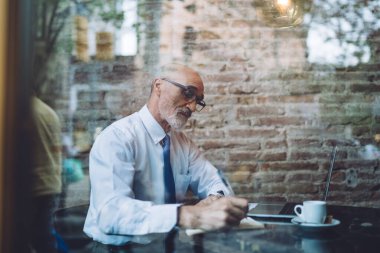 Gözlüklü, olgun, odaklanmış kel bir adamın resmi kıyafetlerini incele. Kafede dizüstü bilgisayarını incelerken not al.