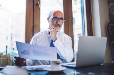 Elinde çizelgelerle kağıt çarşaf tutan ve elinde kahve bardağıyla masada otururken dizüstü bilgisayara bakan yaşlı CEO.