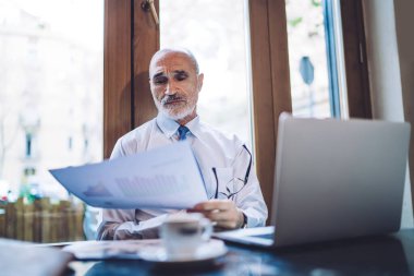 Gözlüklü yaşlı yönetim kurulu başkanı çizelgelerle kağıt çarşafa bakıyor ve pencerenin yanındaki kahve fincanıyla masada otururken dizüstü bilgisayarla çalışıyor.
