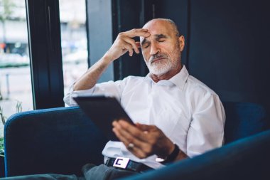 Hüzünlü, kel, resmi kıyafetler giyen, modern tableti karıştıran, gözleri kapalı, elleri kafeteryada oturan bir adam.