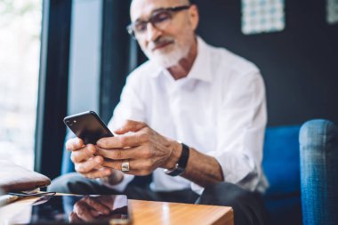 Gözlüklü, mavi sandalyede oturan ve mesaj yazarken akıllı telefon ekranına bakan bulanık yaşlı adam.