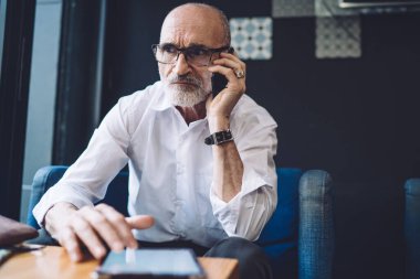 Kel, gri sakallı, gözlüklü, beyaz gömlekli, mavi sandalyede oturan ve telefonla konuşurken yüzünü çeviren yaşlı bir adam. 