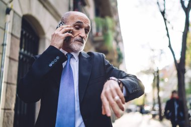 Kendine güvenen, resmi kıyafetli, beyaz sakallı yaşlı bir erkek. Bulanık bina ve ağaçların arka planındaki kol saatine bakarken telefon ediyor. 