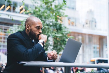 Resmi olarak endişelenen Afrikalı Amerikalı adam dizüstü bilgisayar kullanıyor ve gün ışığında kafeteryada bulanık arka planda düşünüyor.