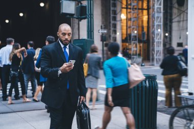 Deri evrak çantası ve mesajlar taşıyan takım elbiseli, kel, Afro-Amerikan bir işadamı şehir merkezinde bulanık arka planda yürürken 