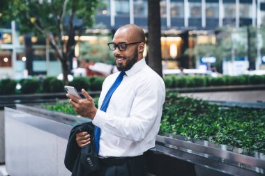 Ofis kıyafetleri ve gözlüklü, takım elbiseli ve modern ofis binasının önünde dikilirken akıllı telefon kullanan kendinden emin Afrikalı Amerikalı bir adam.