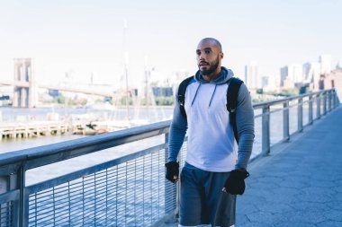 Spor giyimli kel Afrikalı Amerikalı adam modern köprüde yürüyor şehir ve nehir manzaralı arka planda.