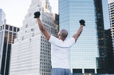 Beyaz tişörtlü, gökdelenlerin yanında el kaldırmış, tanınmayan başarılı etnik atletik adamın arka planı.
