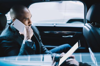 Yoğunlaşmış yönetici Afrika kökenli Amerikalı erkek yönetici cep telefonuyla iş görüşmesi yapıyor ve arabada otururken boş ekranlı tablet kullanıyor.