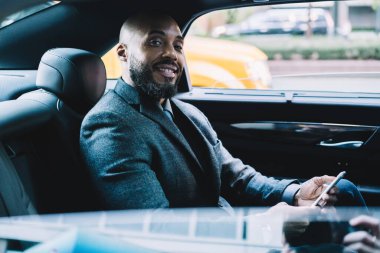 Mutlu sakallı ve kel siyah iş adamı resmi giysiler içinde kameraya bakıyor ve arabada otururken cep telefonunu kullanıyor.