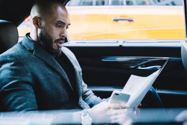 Sıkıntılı profesyonel yönetici sakallı, şık takım elbiseli Afrikalı Amerikalı erkek müdür işe giderken finansal raporları dikkatle okuyor. 