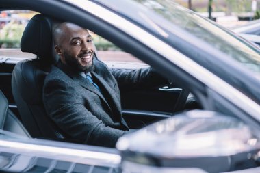 Lüks siyah otomobilleri sürerken şık giyinmiş, kameraya bakıp gülümseyen mutlu Afro-Amerikan bir adamın penceresinden.