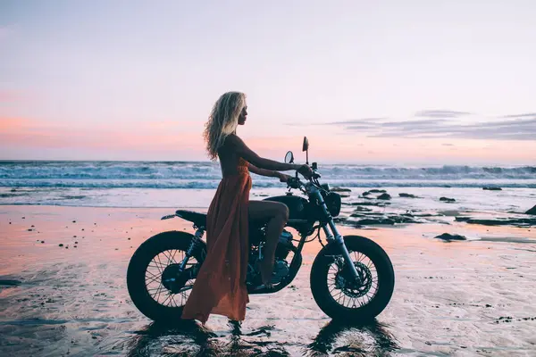 Side view of hot seductive woman in orange light dress opening legs on black motorcycle on seaside on background of breathtaking water and sky