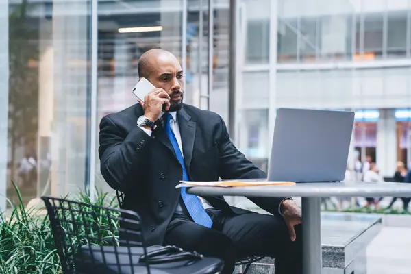 Elegant bald executive black man sitting at table with leather bag on chair near talking on phone while watching laptop in hall of business center