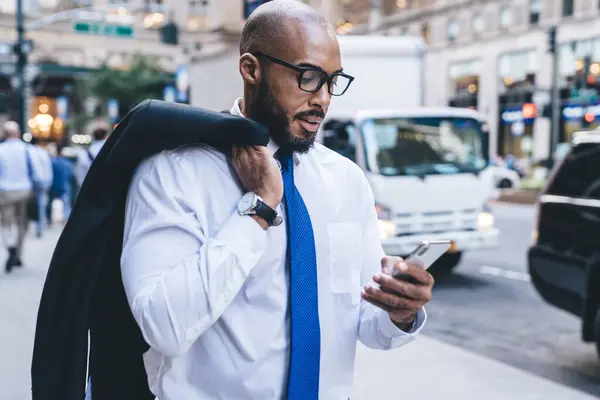 Bald bearded African American businessman in white shirt with blue tie and eyeglasses holding jacket on shoulder standing and messaging on downtown street