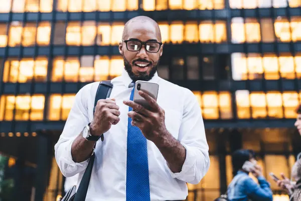 Excited black worker with black beard chatting in social media app and smiling while standing on street in front of glowing windows