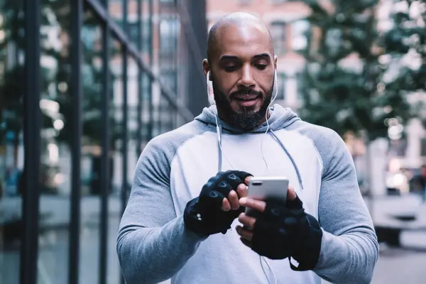African American guy in sportswear listening to music and using smartphone while standing on street near modern office building during outdoor workout