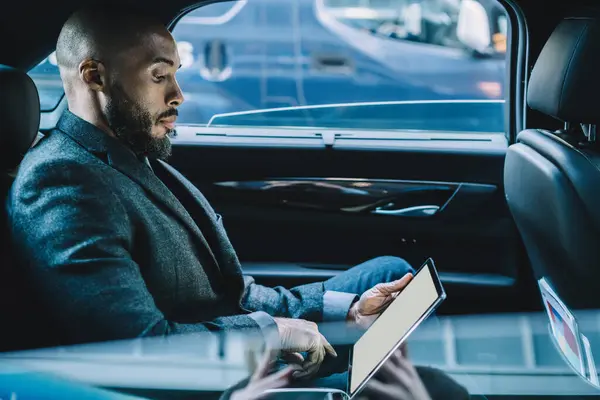 Thoughtful confident ethnic executive man in formal clothes concentrating on screen and interacting with digital tablet with blank screen while sitting in car standing in traffic jam