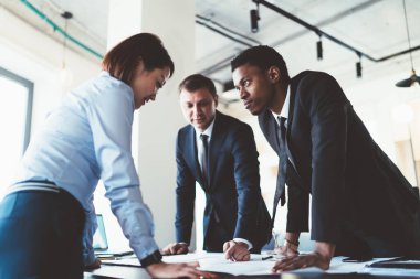 Çok ırklı genç meslektaşlar ellerinde cihazlar ve belgelerle masanın yanında duruyor ve proje planı hakkındaki fikirlerini şirket bürosunun toplantı odasında paylaşıyorlar.