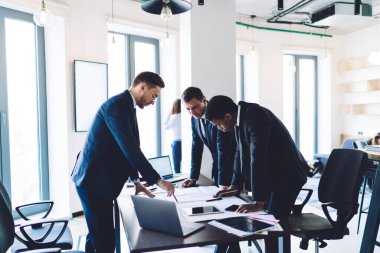 Takım elbiseli, iş projesinden bahseden, fikirlerini paylaşan, ofiste ayakta duran ve yaslanan çeşitli iş arkadaşlarının yan görüntüsü.