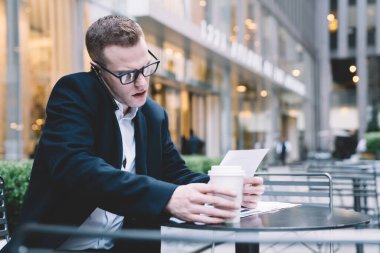 Somurtkan işadamı, siyah ofis takımı ve gözlük takıyor. Sokak kafesindeki masada oturuyor. Telefonu omzuna dayayıp evrak işlerine bakıyor.