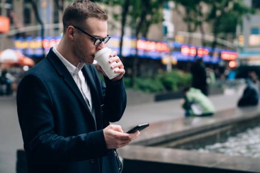 Resmi kıyafetler içinde, gözlüklü ve deri çantalı bir yönetici sokakta dikiliyor ve bulanık arka planda kahve içerken akıllı telefon kullanıyor.