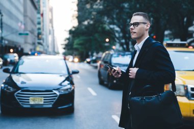 Takım elbiseli, gözlüklü, omuz çantalı genç adam elinde akıllı telefonuyla işlek cadde yolundan karşıya geçerken yan görünüm.